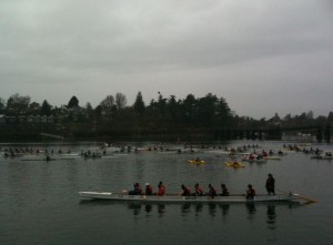 The image shows multiple rowing teams on a body of water, participating in what appears to be a rowing event or race. The sky is overcast, and the surrounding area includes trees and some buildings in the background. The teams are in long, narrow boats, and the rowers are wearing various colors of clothing.