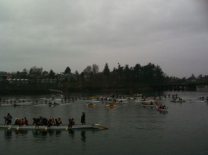 The image shows a group of people participating in a rowing event on a body of water. Several boats with rowers are visible, and the participants appear to be engaged in a race or practice session. The sky is overcast, and there are trees and buildings in the background. The overall atmosphere seems to be calm and focused on the rowing activity.