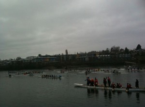 The image shows a cloudy day with a group of people participating in a rowing event on a body of water. Several rowing boats with teams are visible on the water, and there are people standing on a dock or platform near the shore. In the background, there are buildings and trees along the shoreline. The overall atmosphere appears to be that of a community or sporting event.