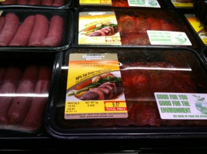 The image shows packages of meat displayed in a refrigerated section of a grocery store. The packages are labeled with information about the contents, including weight and price. One of the labels prominently features a picture of cooked meat with vegetables and text that reads "Good for you, good for the environment." The packages are sealed in plastic and arranged neatly on the shelf.