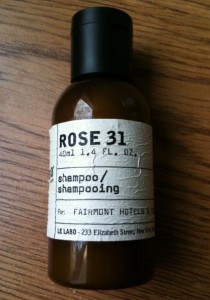 The image shows a small brown bottle with a black cap, containing shampoo. The label on the bottle reads "ROSE 31," "40ml 1.4 fl. oz.," "shampoo/shampooing," and "For: FAIRMONT HOTELS & RESORTS." The bottom of the label includes the text "LE LABO - 233 Elizabeth Street, New York." The bottle is placed on a wooden surface.