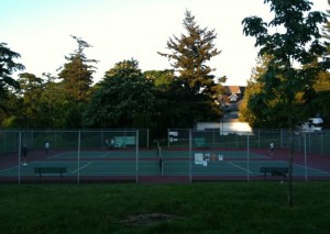 The image shows an outdoor tennis court surrounded by a chain-link fence. There are a few people playing tennis on the court. The court is situated in a park-like area with trees and greenery around it. In the background, there are some buildings partially visible through the trees. The sky is clear, indicating it might be late afternoon or early evening. There are benches outside the fence for spectators or players to sit on.