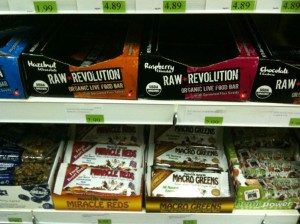 The image shows shelves in a store displaying various health food bars. The top shelf has boxes labeled "Raw Revolution" in different flavors such as Hazelnut, Raspberry, and Chocolate. The middle shelf contains boxes of "Miracle Reds" and "Macro Greens" bars. The price tags are visible above each product.