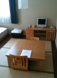 The image shows a small, tidy room with a minimalist design. There is a low wooden table with two chairs, and a piece of paper is placed on the table. In the background, there is a wooden cabinet with a small TV or computer monitor on top. Next to the cabinet, there is a basket with some items in it. A bed is partially visible on the left side of the image, and a window with white curtains and a dark curtain is on the left wall. The floor appears to be covered with tatami mats.