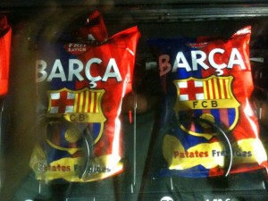 The image shows two bags of potato chips in a vending machine. The bags are branded with the logo and colors of FC Barcelona, a popular football club. The text on the bags reads "BARÇA" and "FCB" with "Patates Fregides" written at the bottom, which is Catalan for "potato chips." The bags are red and blue, matching the team's colors.