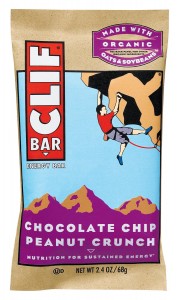 The image shows a package of a Clif Bar. The packaging is primarily beige with a blue background in the center. The Clif Bar logo is prominently displayed on the left side in red and white. The flavor of the bar is "Chocolate Chip Peanut Crunch," which is written at the bottom in white text on a purple background. The package also mentions that it is "Made with Organic Oats & Soybeans" in a purple banner at the top. There is an illustration of a person rock climbing in the center of the package, with mountains in the background. The net weight of the bar is 2.4 oz (68g).