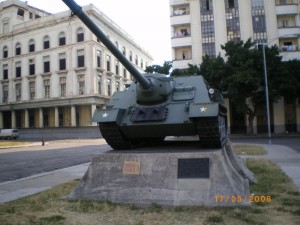 The image shows a military tank displayed on a concrete pedestal in an urban area. The tank is green with a long barrel and has white stars on its sides. The pedestal has plaques attached to it. In the background, there are multi-story buildings and trees. The date on the image is 17/05/2006.