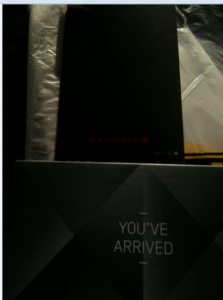 The image shows a dark-colored folder or booklet with the "Air Canada" logo and text on it. Below it, there is a card or sign with the text "YOU'VE ARRIVED" written on it. The background appears to be a plastic or paper surface.