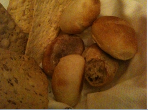 A variety of bread rolls and crackers are placed in a basket lined with a white cloth. The assortment includes round bread rolls, a partially sliced roll, and several types of crackers, some of which appear to have seeds or grains. The lighting is warm and slightly dim.