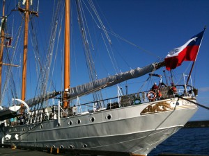 The image shows a large sailing ship docked at a pier. The ship has multiple masts with rigging and a large Chilean flag flying at the stern. The hull of the ship is painted white, and there are people visible on the deck. The sky is clear and blue, indicating a sunny day.