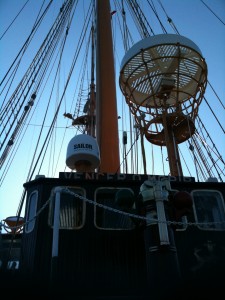 The image shows the upper part of a ship, focusing on the mast and rigging. There are numerous ropes and cables extending from the mast, and a large radar or communication device labeled "SAILOR" is prominently visible. The sky in the background is clear, indicating it is likely daytime. The perspective is from below, looking up at the ship's structure.