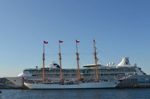 The image shows a large cruise ship docked at a port with a smaller, older-style sailing ship with multiple masts in the foreground. The sky is clear and blue, indicating a sunny day. The sailing ship has several red flags on its masts. The cruise ship is significantly larger and more modern in design compared to the sailing ship.