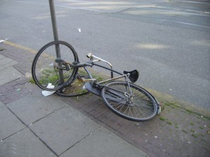 A bicycle is lying on the ground next to a sidewalk. The front wheel is still attached to a pole, while the rest of the bike is detached and lying on its side. The front wheel appears to be bent. The background shows a street with some faint markings.