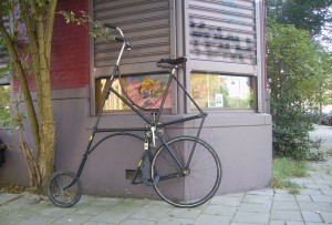 A tall, uniquely designed bicycle is leaning against the exterior wall of a building. The bicycle has an unusually high seat and handlebars, with a small front wheel and a larger rear wheel. The building has a window with some artwork visible inside, and there is a tree to the left of the bicycle. The scene is set on a paved sidewalk with some greenery in the background.