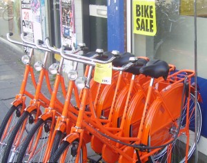 The image shows a row of bright orange bicycles parked outside a shop. There is a sign on the shop window that reads "BIKE SALE." The bicycles are lined up closely together, and there are various posters and advertisements visible on the wall behind them.