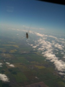 The image shows an aerial view of a landscape with fields and scattered clouds. There is a blurry insect on the window, which is in focus and appears to be flying outside, but is actually on the glass. The horizon is visible in the distance under a clear blue sky.