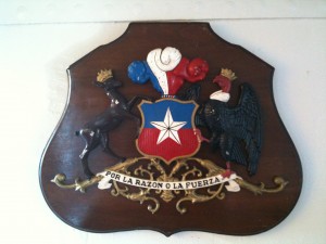 The image shows a wooden plaque featuring a coat of arms. The coat of arms includes a shield with a white star on a blue background, bordered by red. Above the shield are three feathers in blue, white, and red. To the left of the shield is a crowned deer, and to the right is a crowned bird, possibly a condor. Below the shield is a banner with the phrase "POR LA RAZON O LA FUERZA," which translates to "By reason or by force."