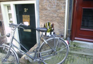 A bicycle is parked in front of a brick building. The bicycle has several empty wine bottles placed on its rear rack. The building has a dark green door with the number "2a" on it, and there is a small window to the left of the door. The ground is paved with cobblestones.