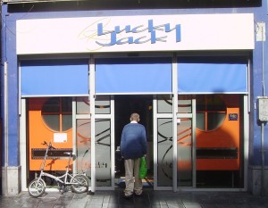 A person is standing in front of the entrance to a store named "Lucky Jack." The store has a blue awning and a sign with the store's name in stylized white and blue letters. The entrance features glass doors with orange and black designs. A bicycle is parked to the left of the entrance. The person is facing the door and appears to be about to enter the store.