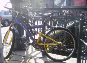 A bicycle with a blue and yellow frame is locked to a metal railing near a staircase leading underground. The background shows a street scene with a few people walking and a parked car. The area appears to be in an urban setting with buildings and other infrastructure visible.