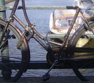 A rusty bicycle is locked to a metal railing by a canal. In the background, there is a boat on the water. The bicycle appears to be old and weathered, with visible rust on the frame and chain. The scene suggests an urban setting near a body of water.