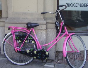 A bright pink bicycle is parked on a sidewalk next to a building. The bicycle has a black seat and black floral-patterned covers on the rear wheel and chain guard. The background includes a stone wall and a glass window with the word "BIKKEMBER" partially visible.