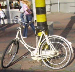 A white bicycle is locked to a black and yellow striped pole on a sidewalk. The rear wheel of the bicycle appears to be bent. In the background, there are people walking and a building with windows.