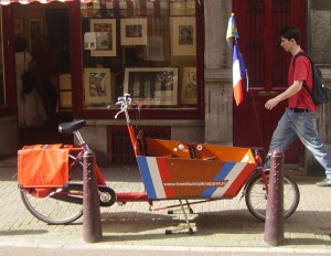 A cargo bike is parked on a street in front of a shop with framed pictures in the window. The bike has a large cargo box painted with red, white, and blue stripes, and a small flag attached to it. A person wearing a red shirt and carrying a backpack is walking past the bike.