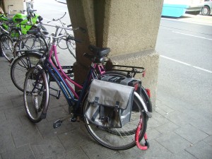 The image shows a bicycle parked on a sidewalk next to a large concrete pillar. The bicycle has a gray pannier bag attached to the rear rack. There are other bicycles parked nearby, and a road with vehicles is visible in the background.