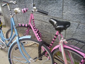 The image shows two bicycles parked next to each other against a stone wall. One bicycle is primarily blue with some black and white striped patterns, while the other is primarily pink with black striped patterns. Both bicycles have black seats and are equipped with various accessories such as bells and racks.