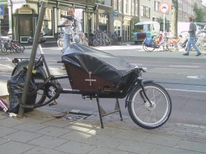 The image shows a cargo bike parked on a sidewalk in an urban area. The bike has a large cargo box at the front, which is covered with a black tarp. The bike is secured to a pole with a lock. In the background, there are several other bicycles parked, a few pedestrians walking, and buildings lining the street. The scene appears to be in a city with a bike-friendly environment.