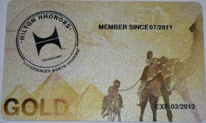 The image shows a Hilton HHonors Gold membership card. The card features the Hilton HHonors logo on the left side with the text "Hilton HHonors" and "Experiences Worth Sharing" around it. The card indicates that the member has been a member since July 2011. There is an expiration date of March 2013 on the bottom right. The background of the card includes an illustration of two people riding camels in a desert-like setting with mountains in the background. The word "GOLD" is prominently displayed in large letters on the bottom left.