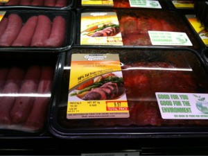 The image shows packages of meat displayed in a refrigerated section of a grocery store. The packages have labels with images of cooked meat and text. One of the labels reads "Good for you, good for the environment." The meat is packaged in black plastic trays with clear plastic covers.