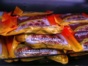 The image shows several packages of kangaroo mince meat stacked on a shelf. The packaging is predominantly yellow with some red and white text. The label on the front of the packages reads "Kangaroo Mince" and indicates a weight of 1 kg.