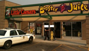 The image shows the exterior of a building with two adjacent businesses: Little Caesars and Booster Juice. A police car is parked in front of the building, and two people are standing near the entrance. The Little Caesars sign is on the left, and the Booster Juice sign is on the right. The storefronts have large glass windows and doors.