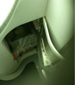 The image shows an open compartment on an airplane, likely in the lavatory area. The compartment appears to contain some plastic bags and other items. The surrounding area includes parts of the airplane's interior, such as walls and flooring.