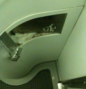 The image shows a damaged and poorly maintained restroom area, likely on a train or airplane. There is a broken panel with exposed tissue paper and a piece of duct tape holding part of the structure together. The floor has a textured mat, and there is a sign with a wheelchair symbol partially visible. The overall condition appears to be unclean and in need of repair.