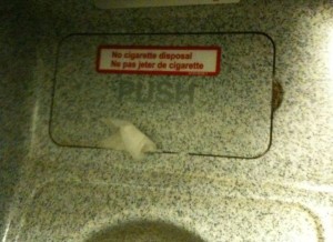 The image shows a metal disposal bin with a push flap. There is a sign on the flap that reads "No cigarette disposal" in both English and French ("Ne pas jeter de cigarette"). A small piece of tissue or paper is stuck in the flap. The surface around the bin is speckled with a gray and white pattern.