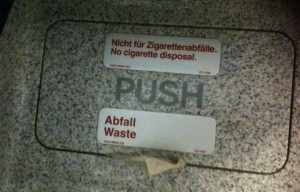 The image shows a waste disposal bin with two signs on it. The top sign reads "Nicht für Zigarettenabfälle. No cigarette disposal." in both German and English. The bottom sign reads "Abfall Waste," also in both German and English. The word "PUSH" is visible in the center of the bin's door. The bin appears to be made of a speckled material.