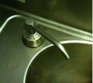 The image shows a close-up of a stainless steel sink with a soap dispenser. The soap dispenser has a long, curved spout and a round base with text on it. The text on the base reads "SOAP PUSH" and "SEIN-DINOS." The sink has a small drain visible at the bottom. The overall setting appears to be a kitchen or bathroom.