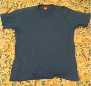 The image shows a blue short-sleeved T-shirt laid flat on a patterned surface. The T-shirt appears to be plain with no visible designs or logos.