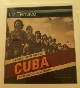 The image is a poster for an exhibition titled "A Revolutionary Project: Cuba from Walker Evans to Now." The poster features a historical photograph of a group of people, possibly revolutionaries, marching together. The text on the poster includes "L2 Terrace" at the top, and the exhibition dates "May 18-October 7, 2011" at the bottom. The word "CUBA" is prominently displayed in large orange letters in the center of the poster.