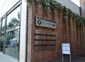 The image shows the exterior of a building with a brick wall adorned with greenery at the top. There is a sign on the wall that reads "RODEO COLLECTION" with a "P" symbol indicating public parking and an arrow pointing to the right. Below this main sign, there are several smaller signs listing various businesses or locations. To the right of the brick wall, there is a white sign with black text that reads "Parking Is Closed." The building has large glass windows, and there are palm trees visible in the reflection.