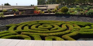 The image shows a beautifully manicured garden with a circular hedge maze design. The hedges are neatly trimmed and form intricate patterns. Surrounding the maze is a stone pathway, and in the background, there are various plants, flowers, and trees. The garden appears to be well-maintained and is likely part of a larger landscaped area.