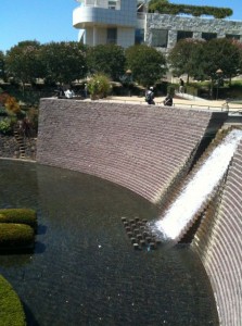 The image shows a landscaped area with a large, curved stone wall that has a waterfall cascading down into a shallow pool below. The pool has neatly trimmed, circular bushes and greenery around it. In the background, there are trees and a modern building with a rooftop garden. A few people are visible near the top of the waterfall, enjoying the scenery. The sky is clear and blue, indicating a sunny day.