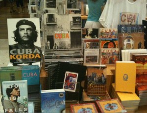 The image shows a display of various books, magazines, and souvenirs related to Cuba. Prominently featured is a large black-and-white poster of Che Guevara with the text "CUBA KORDA." Other items include books with titles such as "Before Night Falls" and "Cuba," as well as postcards, notebooks, and a yellow book with an emblem on the cover. The display appears to be in a store or a market, with a white T-shirt hanging in the background.