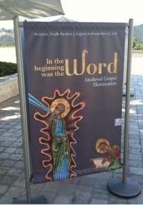 A vertical banner is displayed outdoors on a paved surface. The banner features medieval-style artwork depicting religious figures. The text on the banner reads: "In the beginning was the Word. Medieval Gospel Illumination." Additional text at the top of the banner indicates the location and dates of the exhibition: "Museum, North Pavilion | August 30 - November 27, 2011." The background includes a scenic view with hills and greenery.