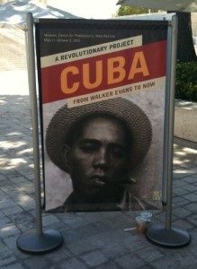 The image shows a poster displayed on a stand outdoors. The poster is titled "A Revolutionary Project: CUBA From Walker Evans to Now." It features a black and white photograph of a person wearing a hat and smoking a cigar. The background of the poster is red and yellow. There is a small cup with a straw placed on the ground next to the stand.