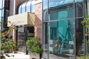 The image shows the exterior of a building with large glass windows. On the left side, there is a storefront with a beige awning that has text on it. There are plants in front of the store, including a flowering bush. On the right side, through the glass window, a person can be seen standing on a ladder, possibly cleaning or performing maintenance. The building has a brick facade and the sky is reflected in the windows.