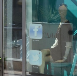 The image shows a store window display with a mannequin dressed in a black and white outfit. The window has stickers for social media platforms Facebook and Twitter, with their respective logos and names. The reflection of the street and surrounding buildings can be seen in the glass.